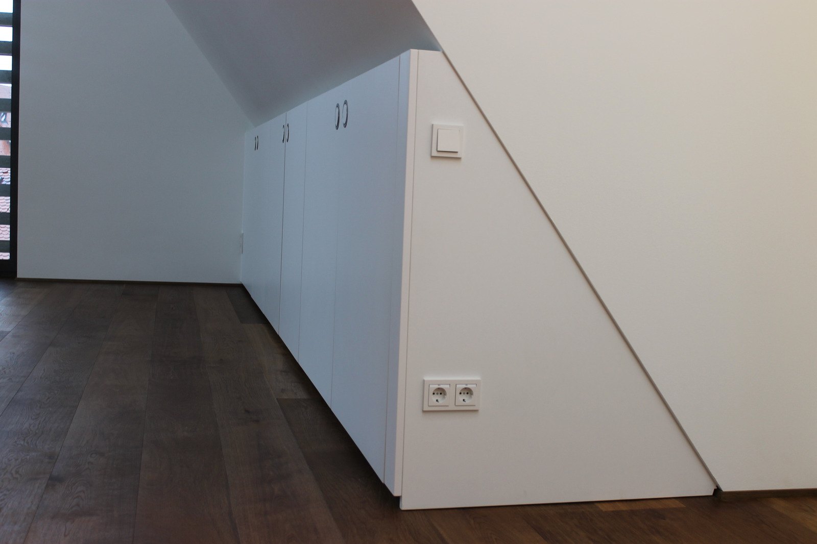 Weißer Einbauschrank entlang einer Dachschräge, rechts die schräg zugeschnittene Seitenwand, davor dunkler Holzboden