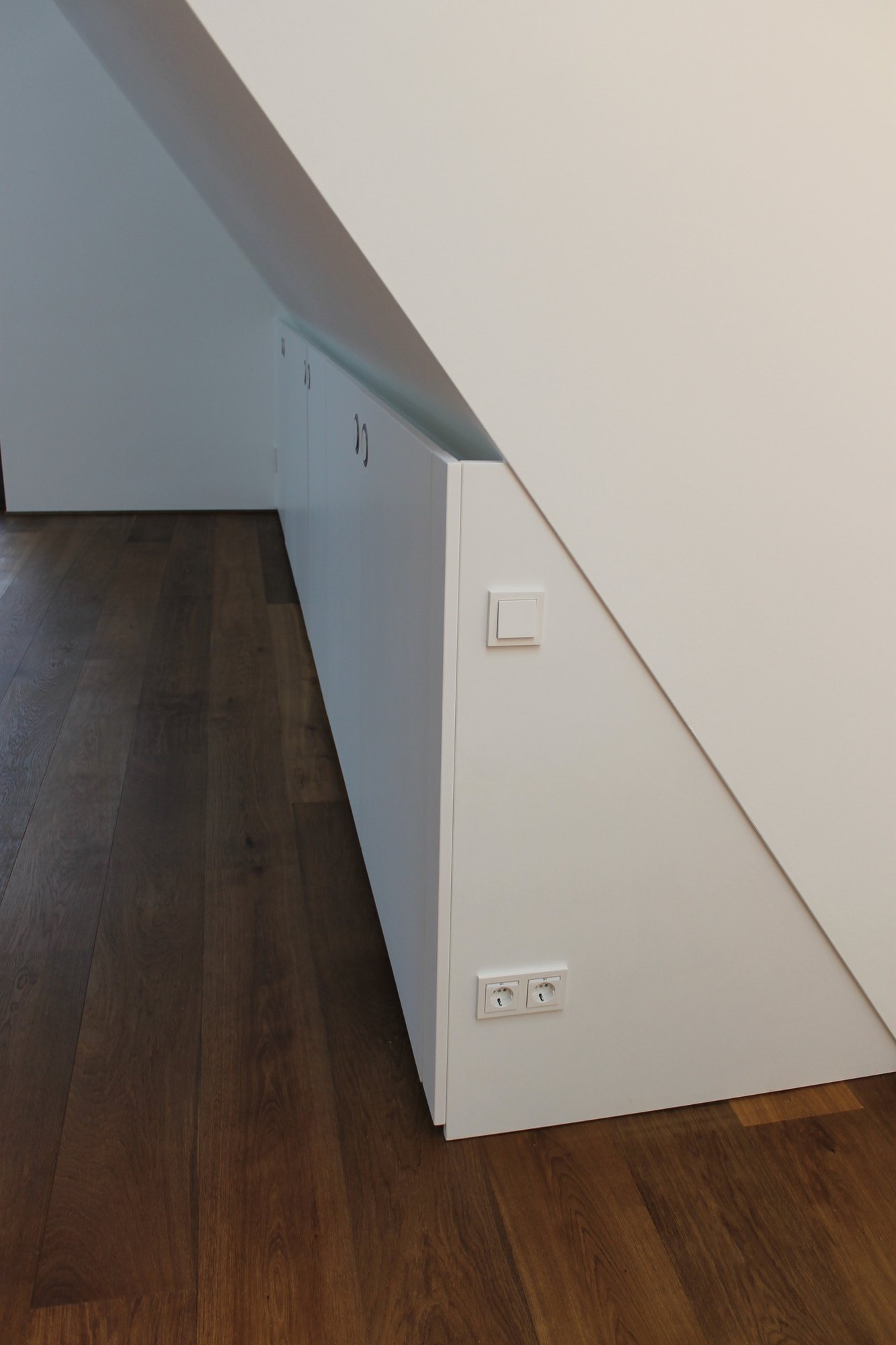 Weißer Einbauschrank unter einer Dachschräge, die Seitenwand folgt der Schräge, davor dunkler Holzboden